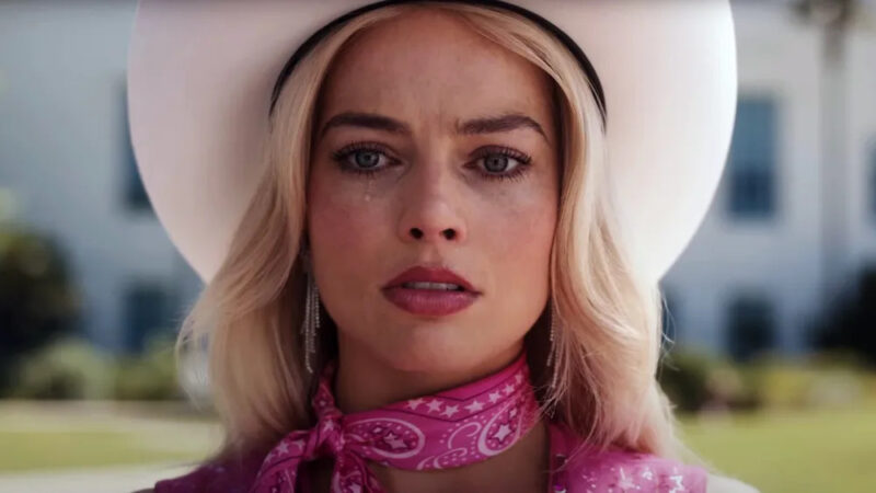 A foto é uma cena do filme Barbie. Nela, temos a atriz Margot Robbie centralizada. Ela é loira, tem olhos claros e usa um chapéu branco de cowboy, além de uma bandana rosa amarrada no pescoço e um brinco prata. Ela está com uma feição séria e uma lágrima escorre do seu olho direito. O frame possui um ângulo fechado, e só é possível ver no fundo desfocado o que aparenta ser um subúrbio.