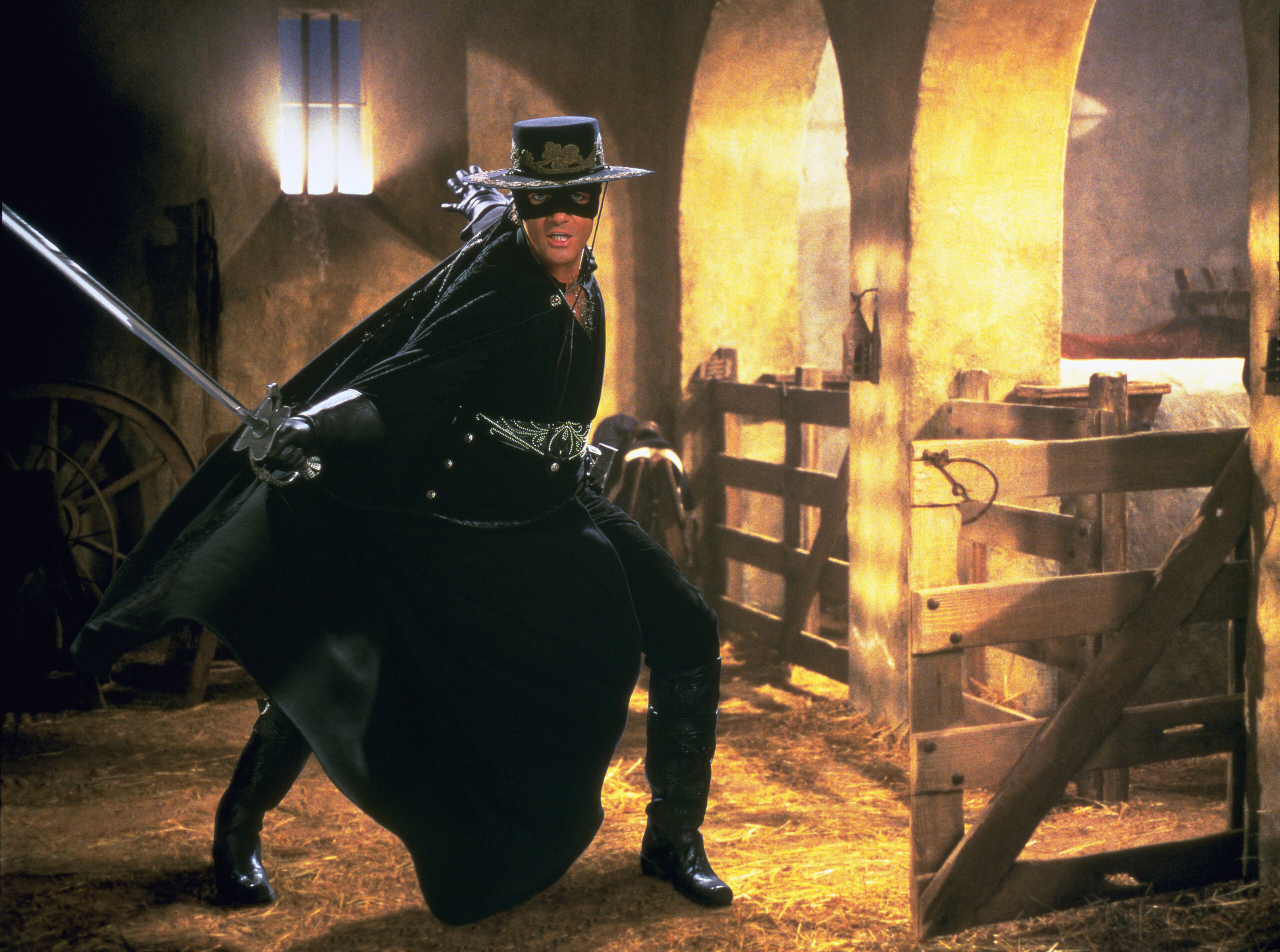 Cena do filme A Máscara do Zorro. Na imagem, o protagonista, Alejandro Murrieta (Antonio Banderas), já atuando como o Zorro, aparece em uma cena de ação com espada rapieira. Murrieta é um homem de pele branca e cabelos e olhos escuros. Ele veste um figurino completamente preto que é acompanhado de luvas e uma capa que se estende até a sola da bota. Ao fundo, o cenário é um celeiro iluminado por cores quentes.