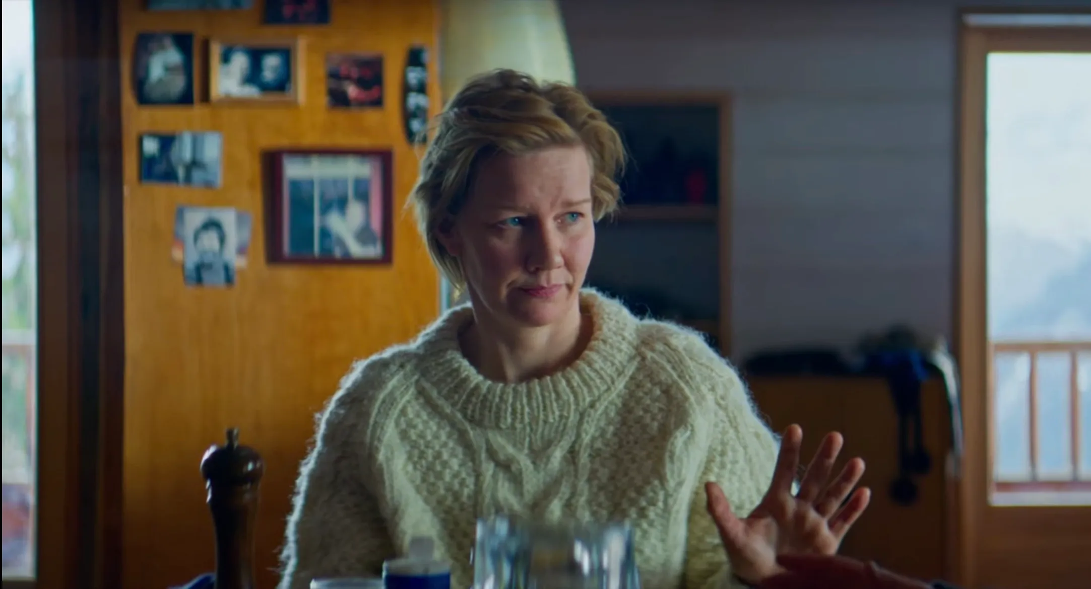 Cena do filme Anatomia de uma queda. Na imagem colorida, a atriz Sandra Huller veste um suéter de cor amarela. Ela está sentada numa mesa na cozinha, com a mão esquerda levemente levantada. Ela é uma mulher branca, loira e de olhos azuis.
