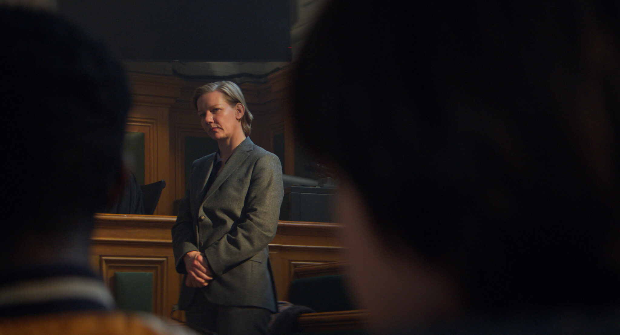 Cena do filme Anatomia de uma queda. Na imagem colorida, a atriz Sandra Huller está num tribunal, vestindo um terno feminino. Ela é uma mulher loira e está com as duas mãos juntas e cruzadas próximo a barriga. Ela está de pé.