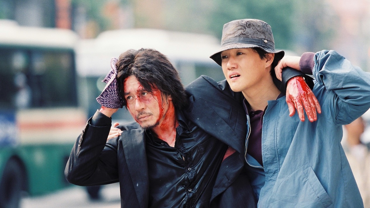 Cena do filme Oldboy. Na imagem, Oh Dae-su (Choi Min-sik) se apoia nos ombros de Lee Woo-jin (Yoo Ji-tae). A câmera os captura a partir da cintura. Oh Dae-su é um homem sul-coreano de cabelos e olhos escuros que segura um pano na cabeça para estancar todo o sangue que escorre pelo terno preto. Lee Woo-jin é um homem sul-coreano de cabelos e olhos escuros que veste uma jaqueta azul fechada por cima de uma blusa roxa, além de um chapéu de pescador na cor cinza. Ao fundo, o cenário é uma rua movimentada pelo tráfego, porém desfocada, deixando os personagens em destaque na composição.