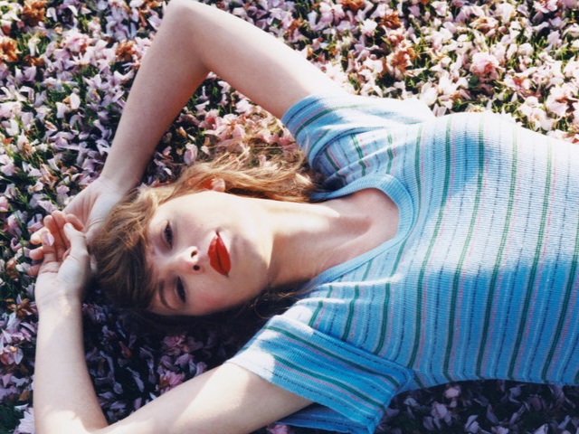 Taylor, uma mulher branca de cabelos loiros e compridos, e batom vermelho, está deitada no chão de grama coberto por flores lilás, com os braços acima da cabeça, usando uma camiseta listrada clara, iluminada por um sol da manhã.