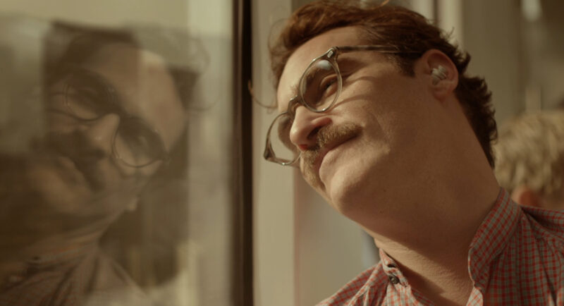 Cena do filme Ela. O personagem principal, Theodore, um homem branco de óculos redondos, cabelo marrom e bigode está olhando para seu reflexo na janela do metrô que tem predominância de tons claros com um leve sorriso.