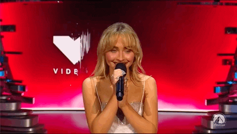 Imagem em movimento de Sabrina Carpenter, mulher branca, falando no microfone no palco do VMAs. A imagem mostra a artista segurando o microfone e olhando em direção à platéia. 