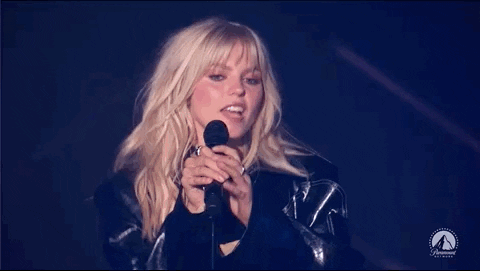 Imagem em movimento de Reneé Rapp, mulher loira, cantando no palco do VMAs. A imagem mostra a cantora, que veste uma jaqueta preta, tirando o microfone do suporte e o levando até a mão. 