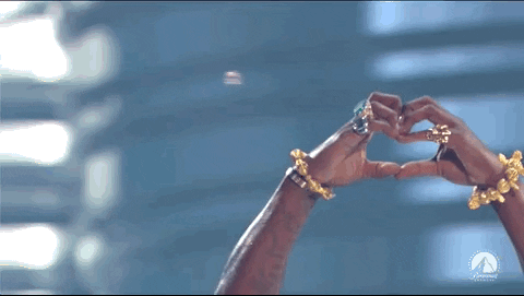 Imagem em movimento do rapper Diddy, homem negro, performando no palco do VMAs. A imagem mostra o artista fazendo um sinal de coração com a mão. Ele usa óculos de sol preto, uma blusa regata vermelha e uma medalha dourada no pescoço.