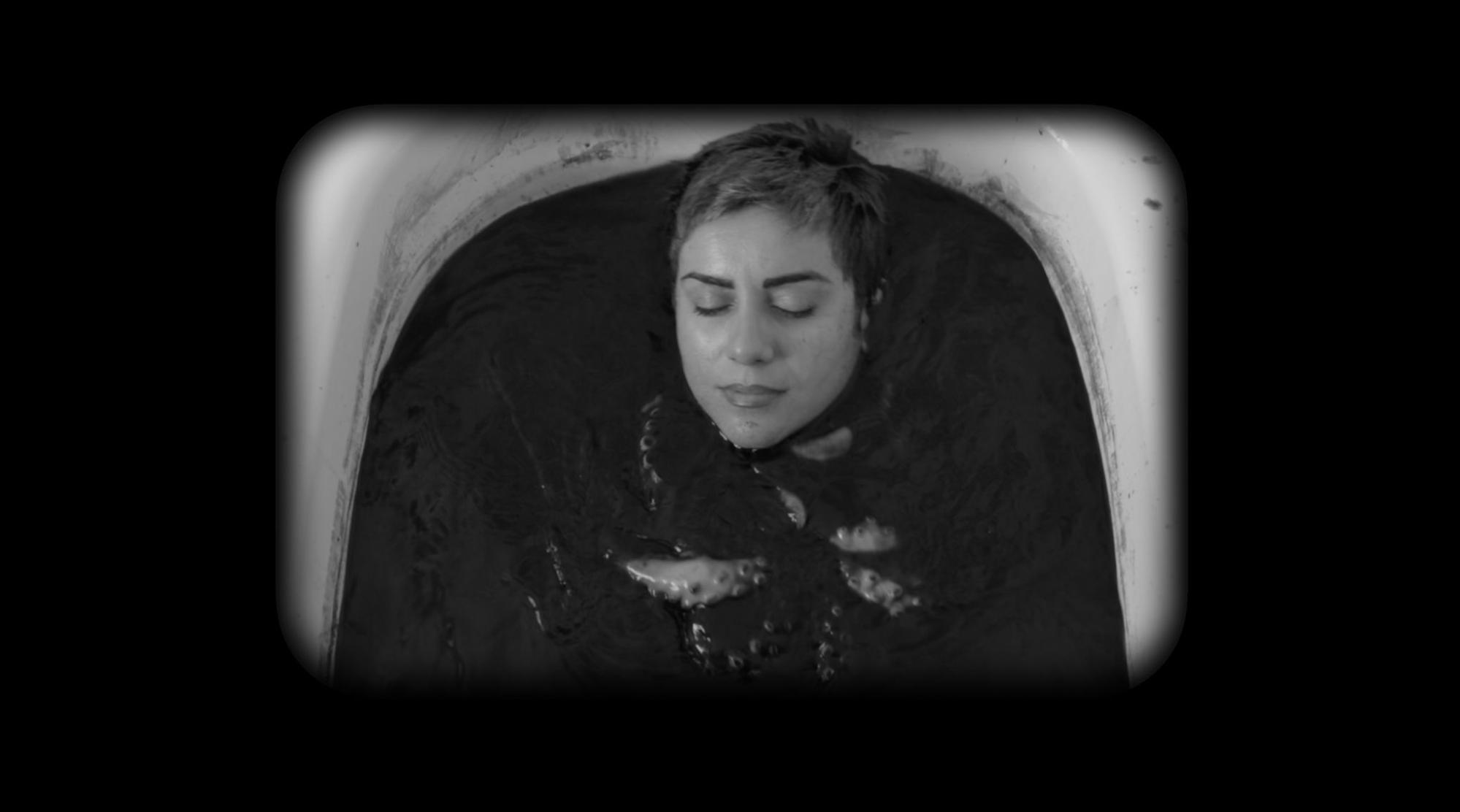  Cena do filme Como você se atreve a desejar algo tão terrível. Na imagem, a cineasta Mania Akbari, uma mulher iraniana de cabelos curtos, aparece em uma banheira. Ela está mergulhada em um líquido escuro com um polvo em seu colo. O retrato está em preto e branco.