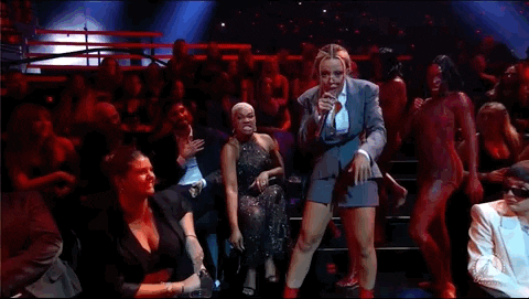  Imagem em movimento de Doja Cat, mulher negra, no palco do VMAs. A imagem mostra a cantora performando na plateia da premiação. Ela usa um óculos arredondado, um terno cinza com uma saia da mesma cor e calça um salto vermelho. 