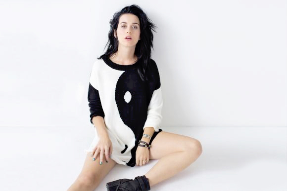  Fotografia da cantora Katy Perry. Na imagem, Perry, uma mulher branca de cabelos escuros e olhos claros, é fotografada sentada em um estúdio de fundo branco. Ela veste um grande suéter de mangas longas com a estampa yin-yang.