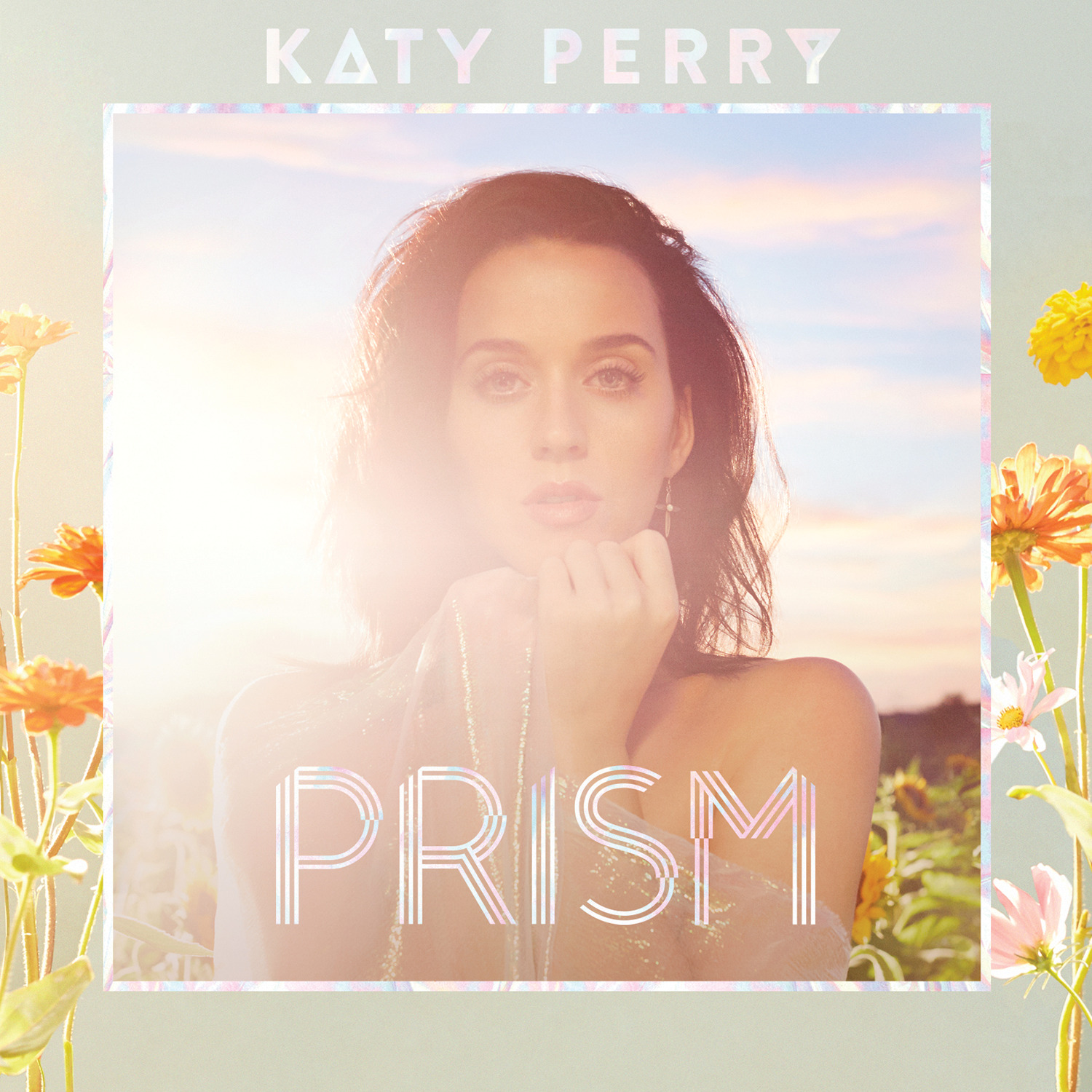 Capa do álbum PRISM da cantora Katy Perry. Na arte da capa, a imagem da cantora está centralizada a partir do busto. Uma borda estilizada apresenta uma paleta de tons suaves com flores nas cores laranja e amarelo. Perry é uma mulher branca de cabelos escuros e olhos claros. Ela está em um campo de girassol. No topo da arte da capa, o nome “Katy Perry” aparece em letras garrafais. Já na parte inferior, o título do disco “PRISM” está escrito da mesma forma.