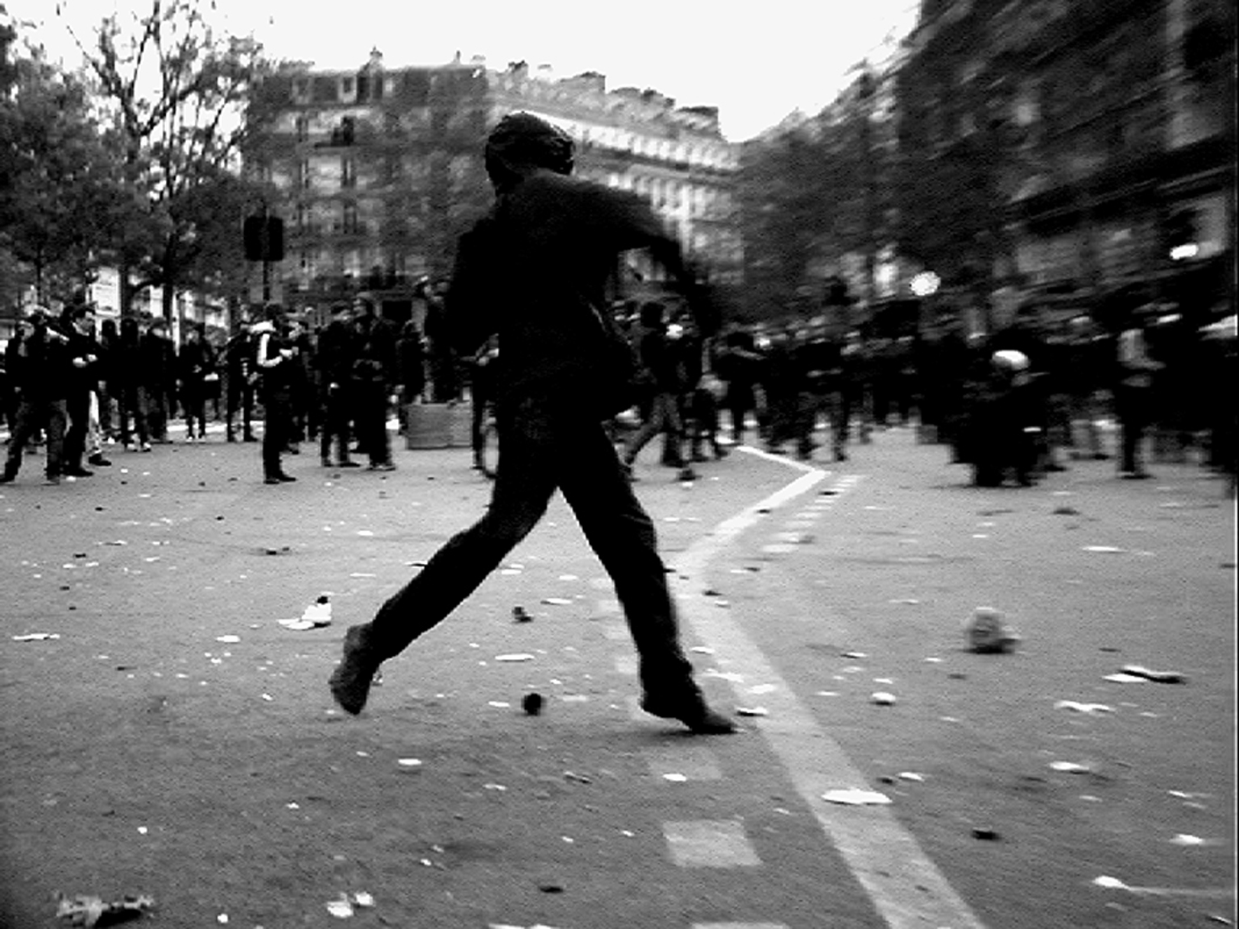 Cena do filme Paris é uma Festa - Um filme em 18 Ondas. A fotografia em preto e branco é um pouco tremida,  e retrata um momento de ação de um manifestante em posição para atirar algo em um protesto nas ruas de Paris. O chão está sujo e, ao fundo, podemos visualizar diversas pessoas pelo centro da cidade. 