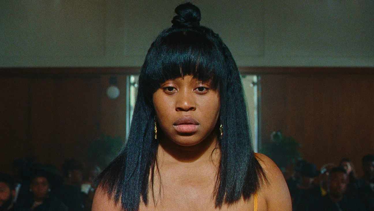 Cena da série Enxame. Imagem da protagonista Dre, centralizada, do tronco para cima. Ela está olhando para frente com uma expressão triste.]