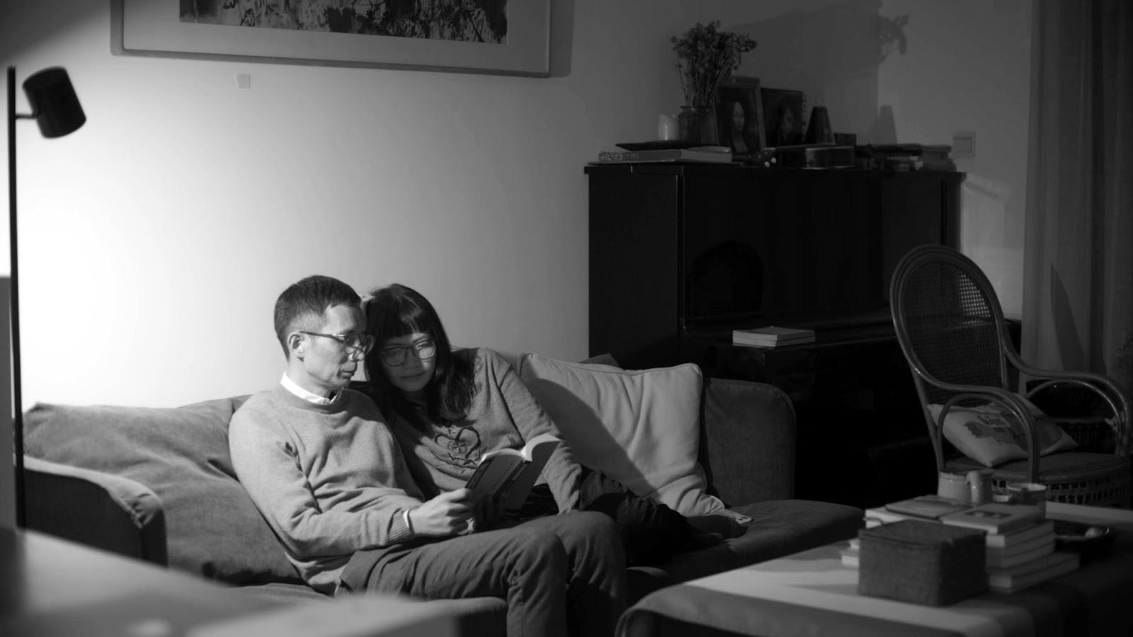Foto de cena do filme Conversas Pela Noite. Na imagem, em preto e branco, está à esquerda, o casal de protagonistas lendo um livro e sentados no sofá. Eles são adultos de meia-idade, chineses e usam óculos. Na frente deles, há uma pequena mesa com livros, à direita um piano e uma cadeira. 