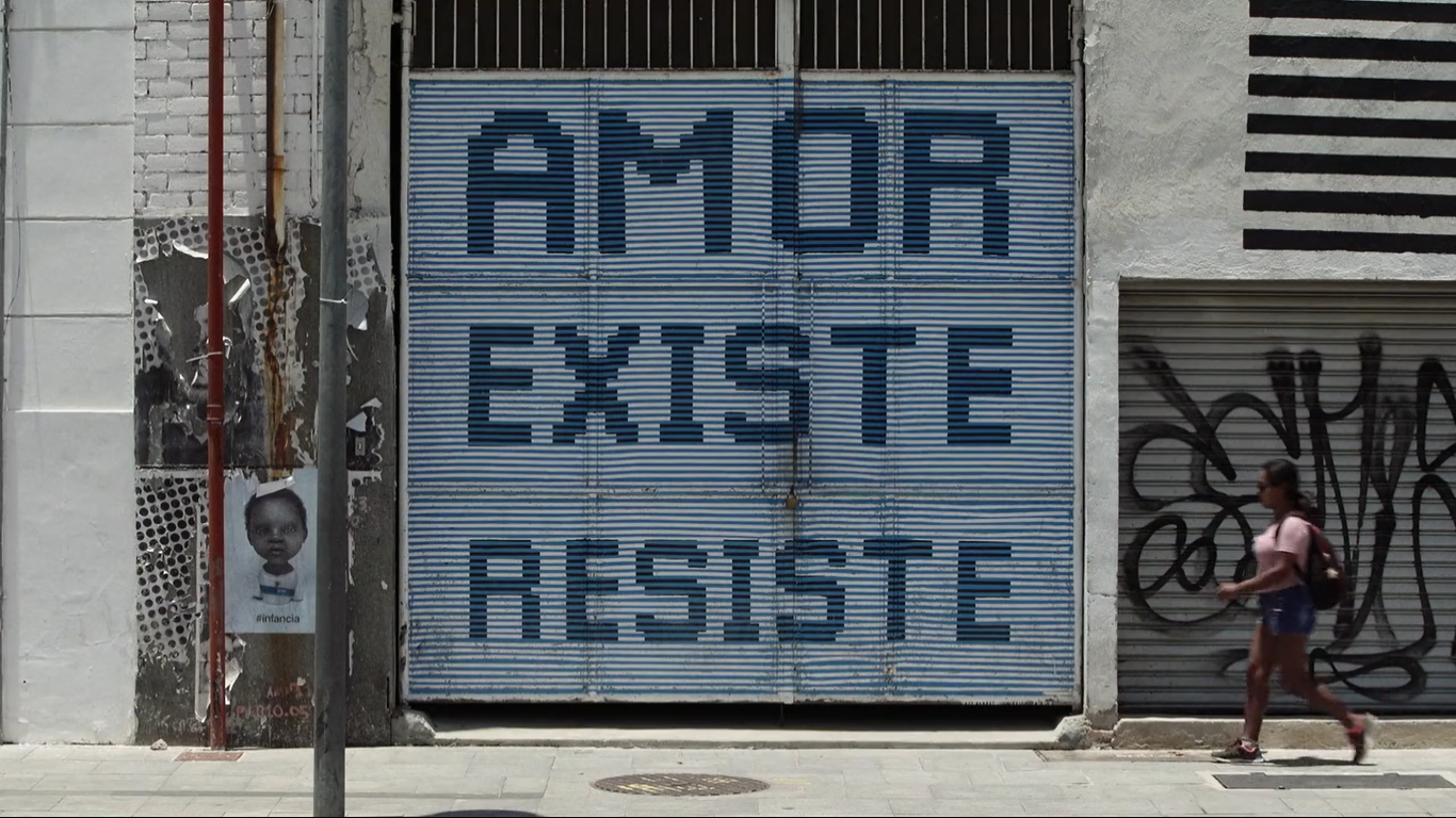 Cena do documentário Pele, de 2021. A imagem mostra uma parede de cor branca, e um portão azul-claro. No portão, há o escrito “amor existe resiste”, em caixa alta, na cor azul-escuro.
