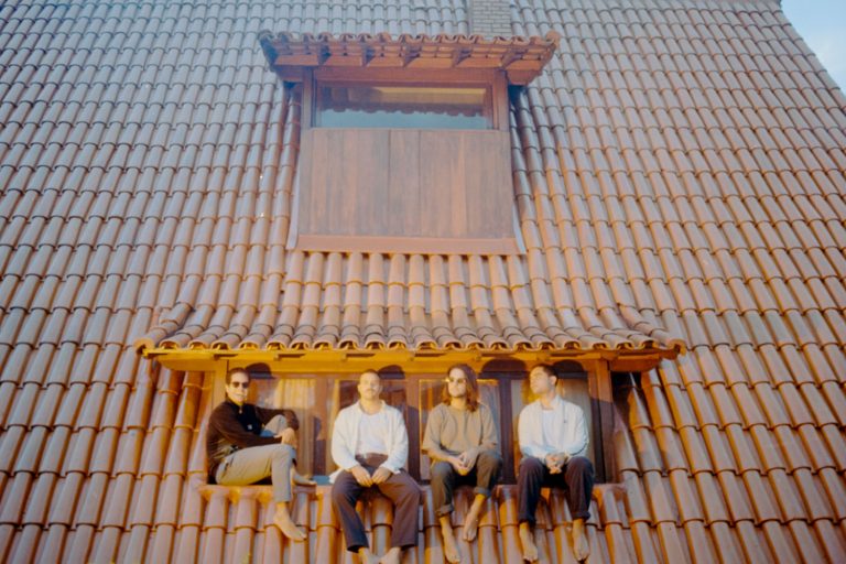 Quatro homens sentados na beira de uma janela presente em um telhado de uma casa. Estão centralizados na fotografia e usam roupas básicas, camisas e calças com paleta de cores terrosa e branca. Está escurecendo e os homens, integrantes da banda, estão sendo iluminados por uma luz amarelada.