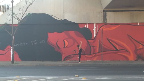 Cena do documentário Pele, de 2021. A imagem mostra um apoio de viaduto, em que a metade inferior possui um grafite de uma mulher deitada, em tons de vermelho, com cabelos pretos. Em frente ao grafite, há uma mulher branca, de cabelos escuros e roupas pretas praticando ioga