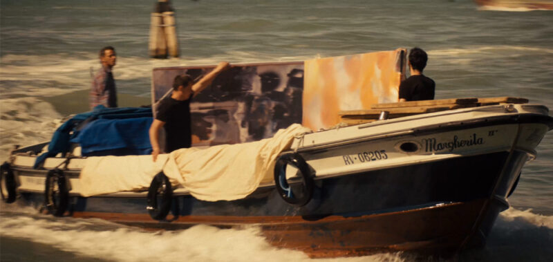 Cena do filme Tomando Veneza. Na imagem há dois homens segurando uma tela pintada à mão nos tons de cinza e amarelo. Os dois estão em uma lancha branca que é conduzida por um terceiro homem. A lancha flutua em um rio esverdeado de Veneza.