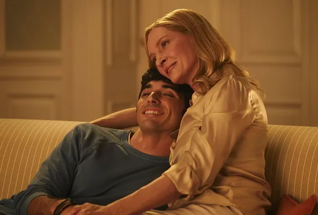 Cena do filme Vermelho, Branco e Sangue Azul, na qual Alex e sua mãe, interpretada por Uma Thurman, estão sentados no sofá da Casa Branca enquanto se abraçam e sorriem. Alex está posicionado à esquerda e sua mãe à direita; ela apoia sua cabeça na de Alex durante o abraço.