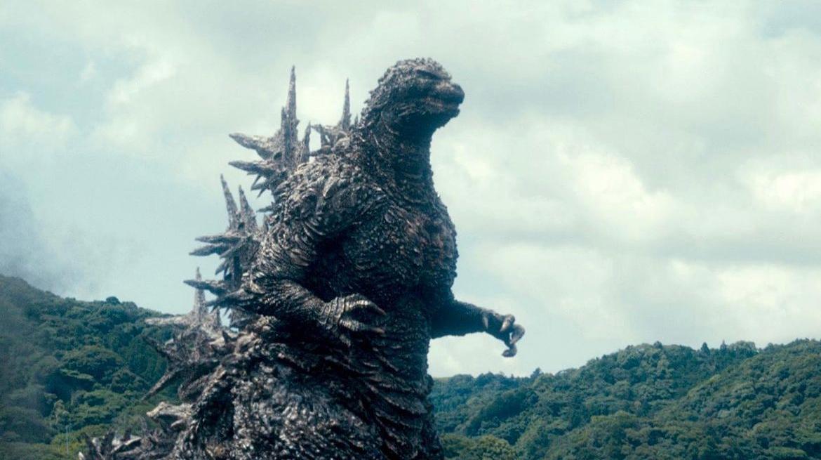 Cena do filme Godzilla Minus One. No centro da imagem aparece o Godzilla, um lagarto que anda em duas patas e possui espinhos nas costas. No fundo da imagem aparece uma floresta e o céu.