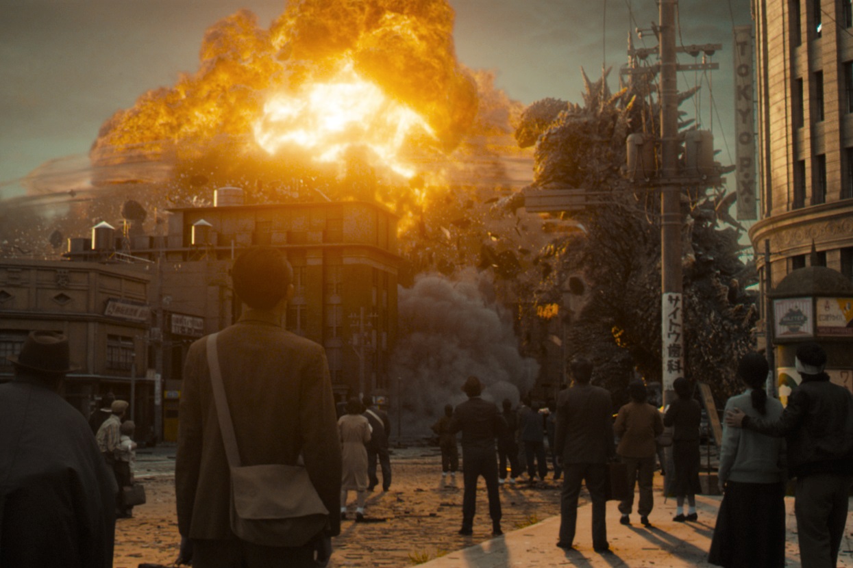 Cena do filme Godzilla Minus One.A imagem é enquadrada na altura dos seres humanos, que observam, atônitos e de costas para a câmera o Godzilla, que está destruindo e explodindo um prédio da cidade. O cenário é Tóquio, e além do Godzilla, as pessoas na imagem enxergam uma explosão causada pelo monstro.