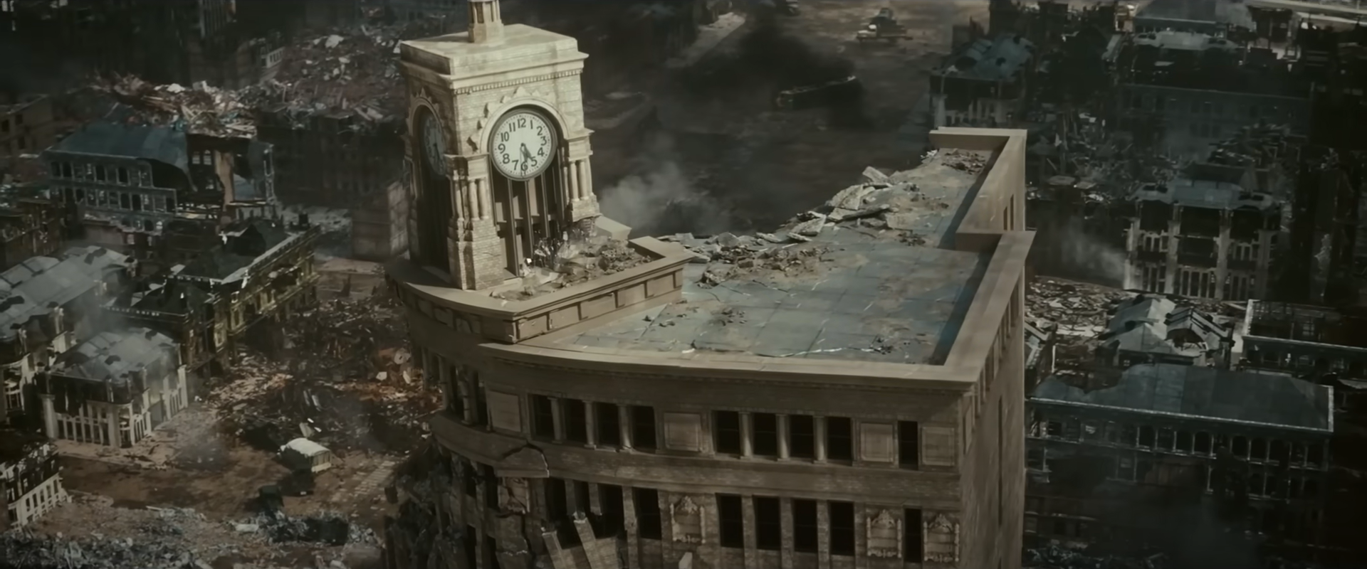 Cena do filme Godzilla Minus One. No centro da imagem aparece um prédio bege destruído, nele tem uma torre com um relógio de ponteiro. No fundo da imagem aparecem escombros de prédios e casas. 
