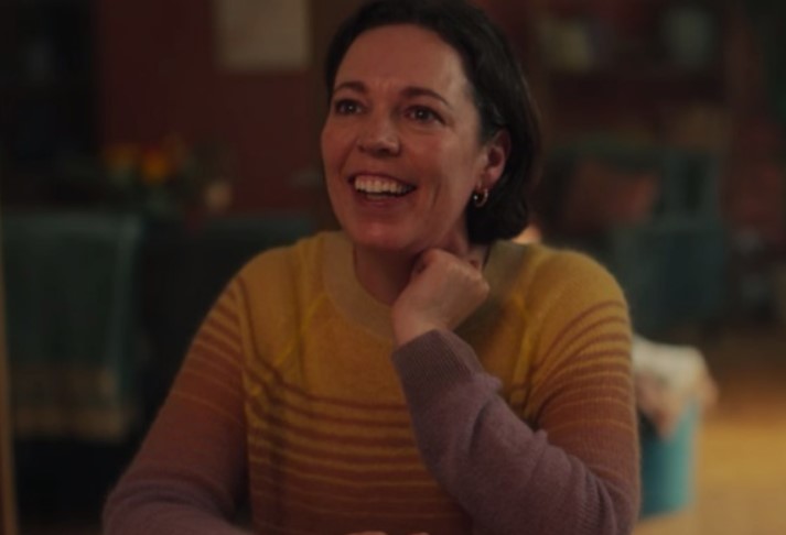 Cena da série Heartstopper. Na imagem, Olivia Colman, uma mulher branca que interpreta Sarah, veste um suéter amarelo com listras vermelhas e manga roxa. Ela está sorrindo enquanto conversa com alguém.
