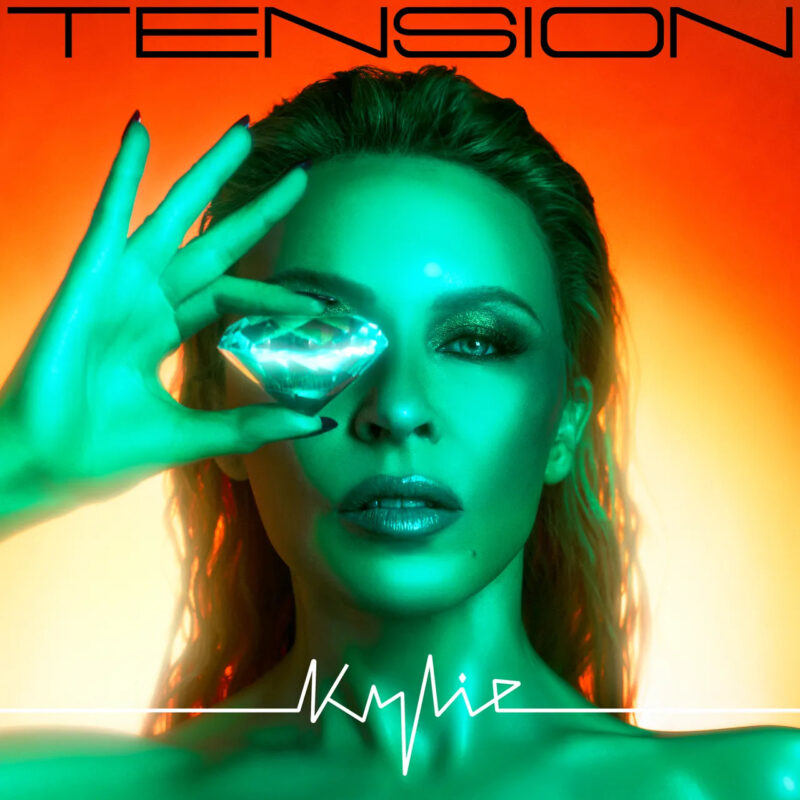 Capa do disco Tension de Kylie Minogue. Na arte de capa, Minogue, uma mulher australiana, branca, de cabelos e olhos claros, encara a câmera enquanto segura um diamante com uma das mãos que, por sua vez, encobre um de seus olhos. Ao fundo, a cor predominante é laranja e uma luz verde encobre a cantora, fotografada dos ombros para cima. Na parte superior da capa, está o título do álbum, Tension, e, na parte inferior, o nome dela, Kylie