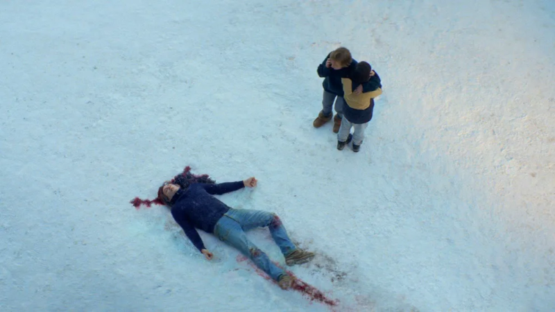 Cena do filme Anatomia de uma Queda. Vista de cima, o chão está coberto de neve, um homem está deitado sobre uma poça de sangue embaixo de sua cabeça. De pé, olhando para ele, uma mulher usa o telefone enquanto abraça um garoto em direção ao seu peito.