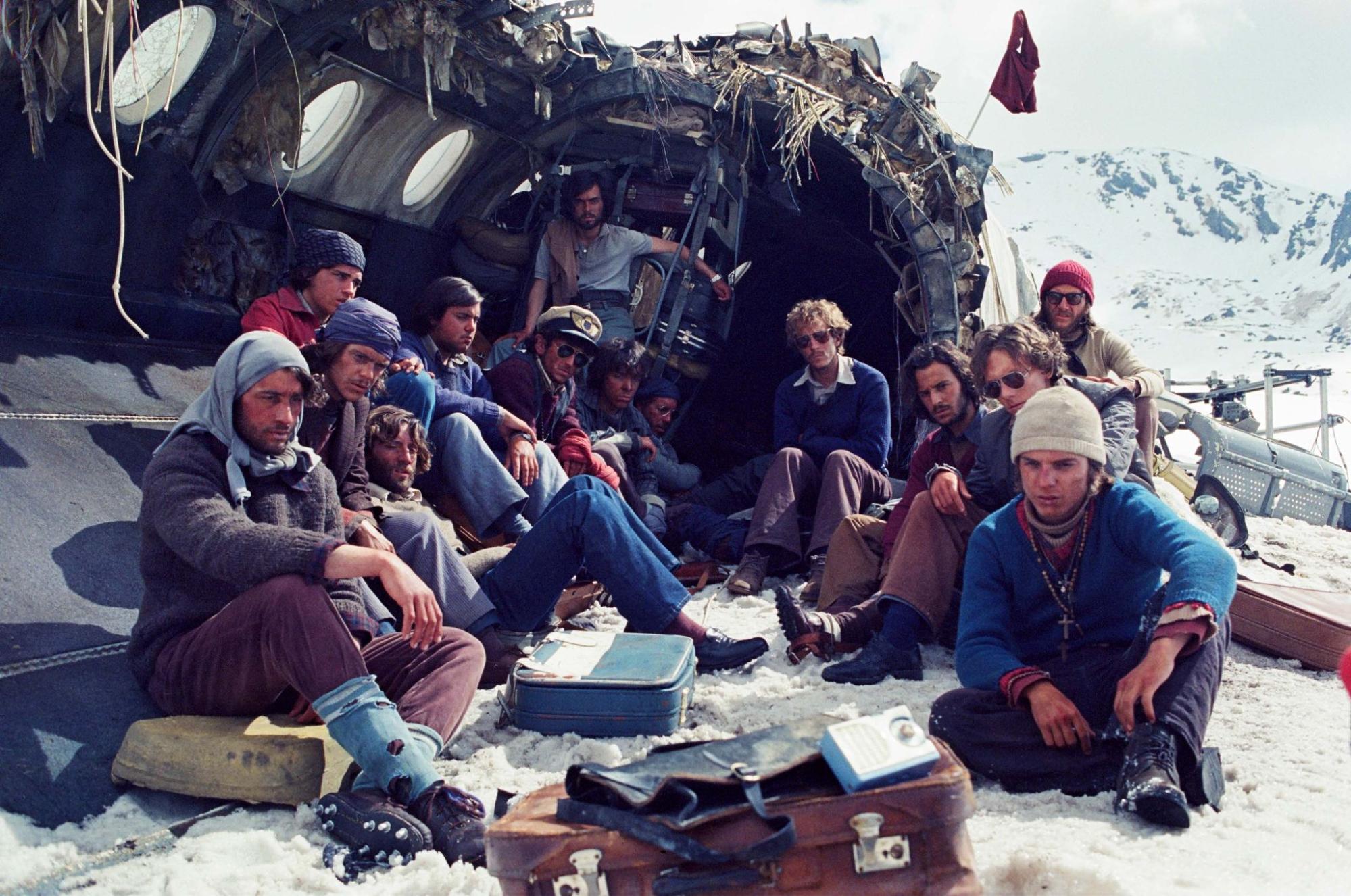 A foto é uma cena do filme A Sociedade da Neve. A foto mostra 14 dos 16 sobreviventes sentados na frente da fuselagem do avião. Estão sentados na neve, alguns em cima da lataria ou de malas. O horizonte atrás do avião mostra as cordilheiras dos Andes