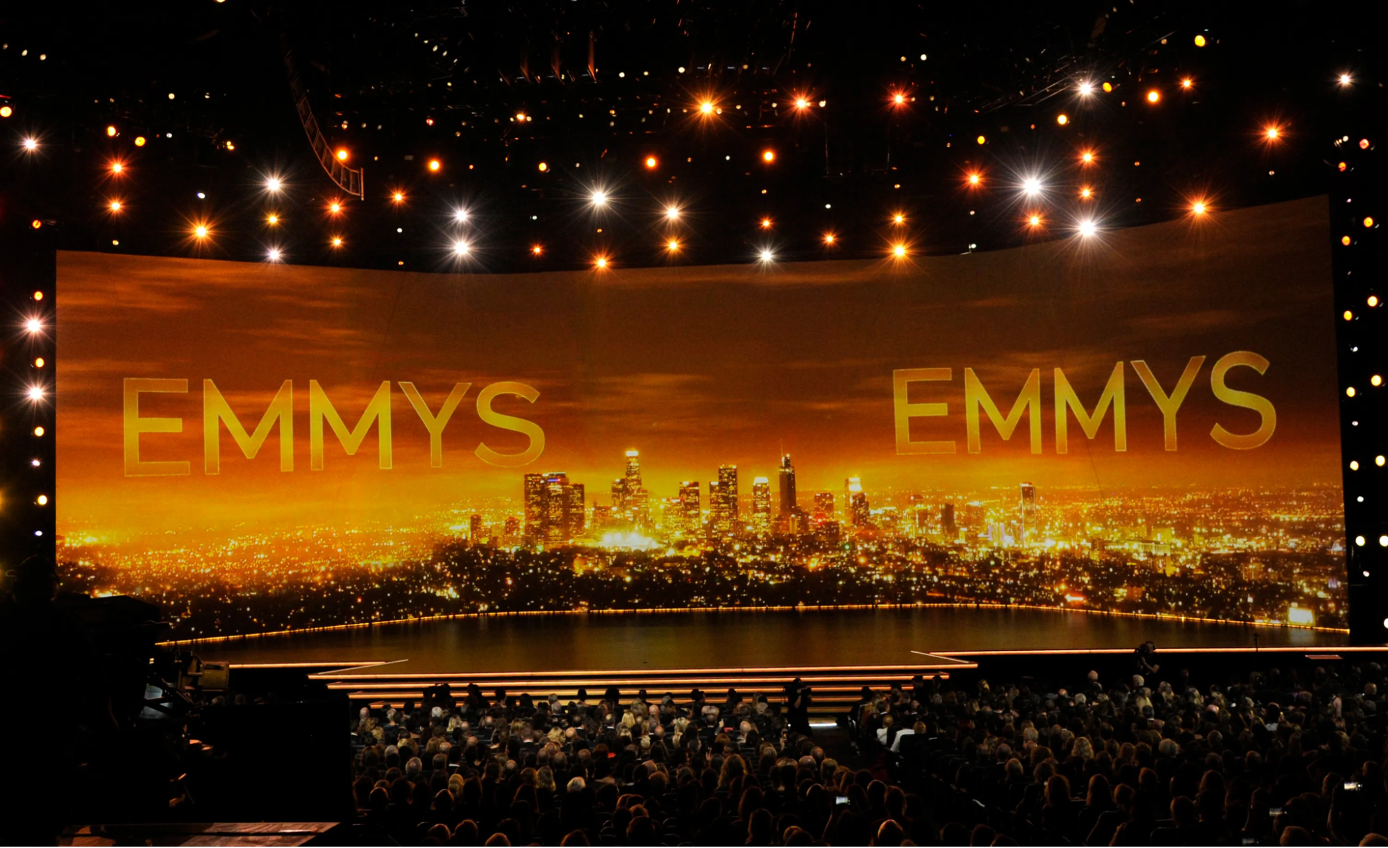 Foto do palco do Emmy de 2021. O cenário da premiação é composto por um telão que mostra os prédios de uma cidade, com um pôr do sol alaranjado se misturando ao letreiro “Emmys”, da mesma cor. Além disso, na plateia há várias pessoas, olhando para o palco.