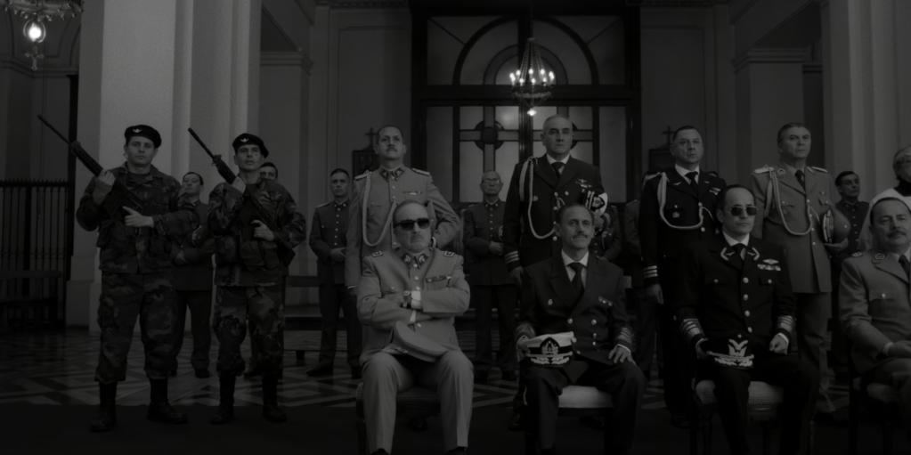 Cena do filme El Conde(2023), Pinochet (Jaime Vadell) aparece no centro da imagem, de terno e óculos escuros, a sua esquerda estão sentados oficiais de alta patente, e a sua esquerda soldados uniformizados empunhando fuzis