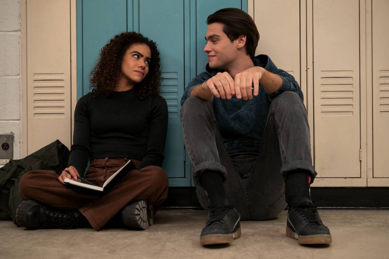 Ginny (Antonia Gentry) sentada no chão, no corredor da Wellsbury High School, ao lado de Marcus (Felix Mallard).]