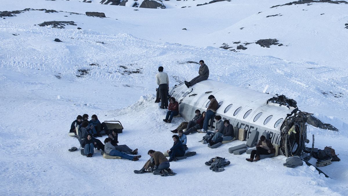  A foto é uma cena do filme A Sociedade da Neve. A foto mostra 20 pessoas ao redor da carcaça do avião, a maioria sentada em casacos ou apoiados no próprio avião. Ao redor, há apenas neve e pedras]
