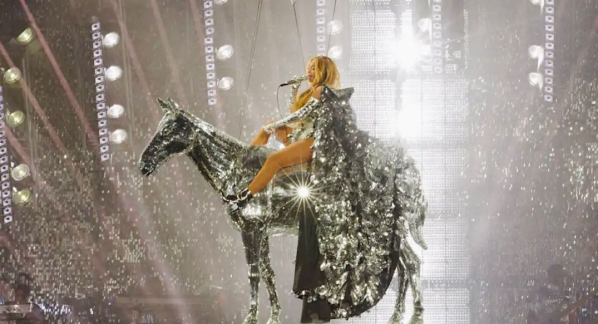 Cena do filme Renaissance: a film by Beyoncé. Na imagem vemos Beyoncé montada em cima de um cavalo feito de metal todo prateado que está sendo erguido por fios de aço. Beyoncé, uma mulher negra com cabelos loiros, usa um vestido prateado com uma imensa cauda, nos pés ela usa botas também prateadas que chegam à altura de seus joelhos. A cantora está segurando um microfone com sua mão direita enquanto canta uma canção. Ao fundo, é possível ver luzes brilhantes e parte dos assentos do estádio.