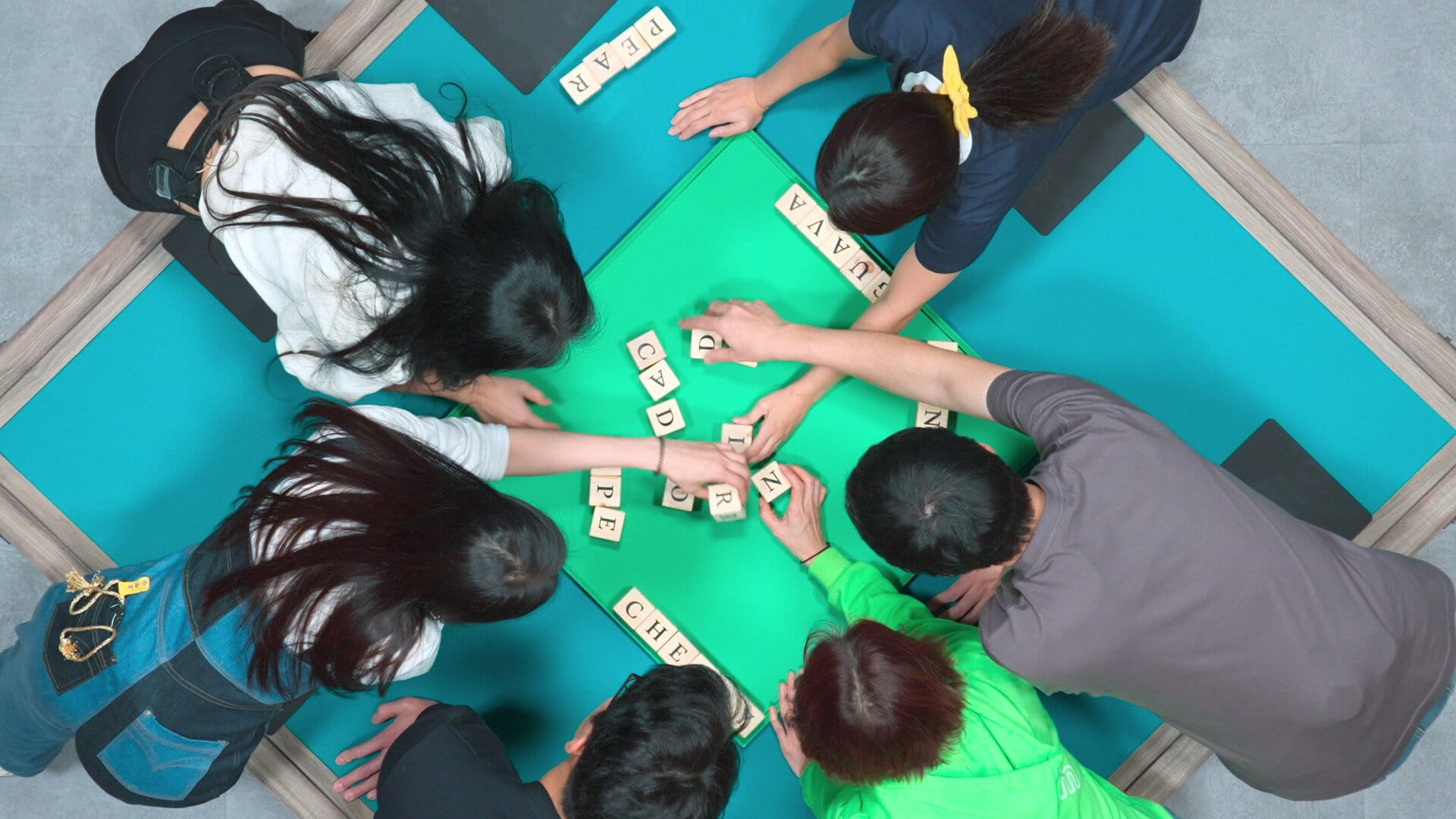 Cena de O Jogo do Diabo. De cima, seis pessoas asiáticas são vistas debruçadas sobre uma grande mesa verde; todos vestem roupas casuais. Estão mexendo em blocos de madeira com diferentes letras.