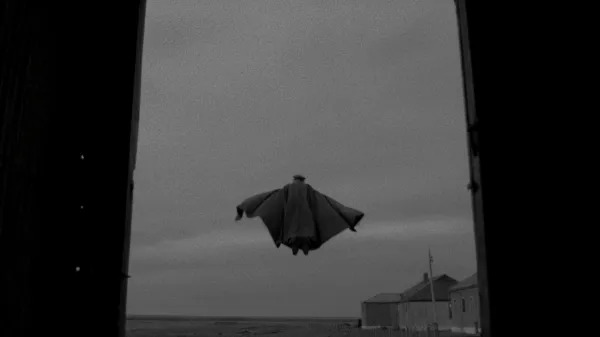  Cena do filme El Conde(2023). Pinochet (Jaime Vadell) voa com sua capa ao vento, e um quepe na cabeça, a imagem é vista da perspectiva de uma porta e no plano há parte da fazenda e um céu acinzentado