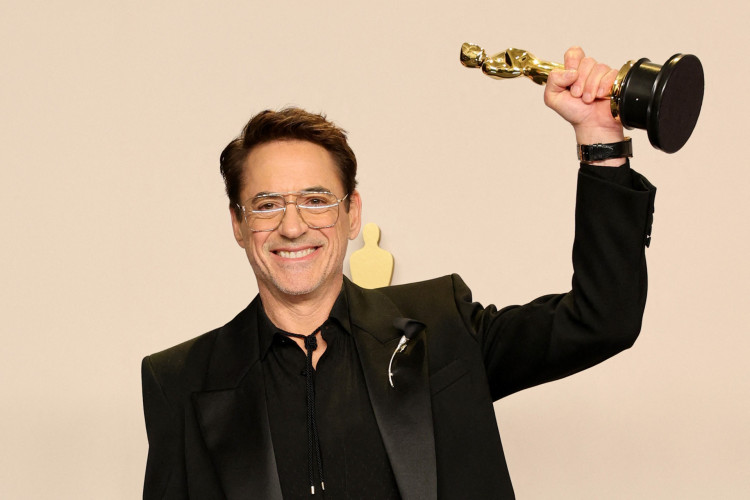 Foto do ator Robert Downey Jr, homem branco de cabelos castanhos. O ator está utilizando um terno preto e segurando a estatueta do Oscar na mão esquerda. O prêmio é representado por uma estatueta dourada.