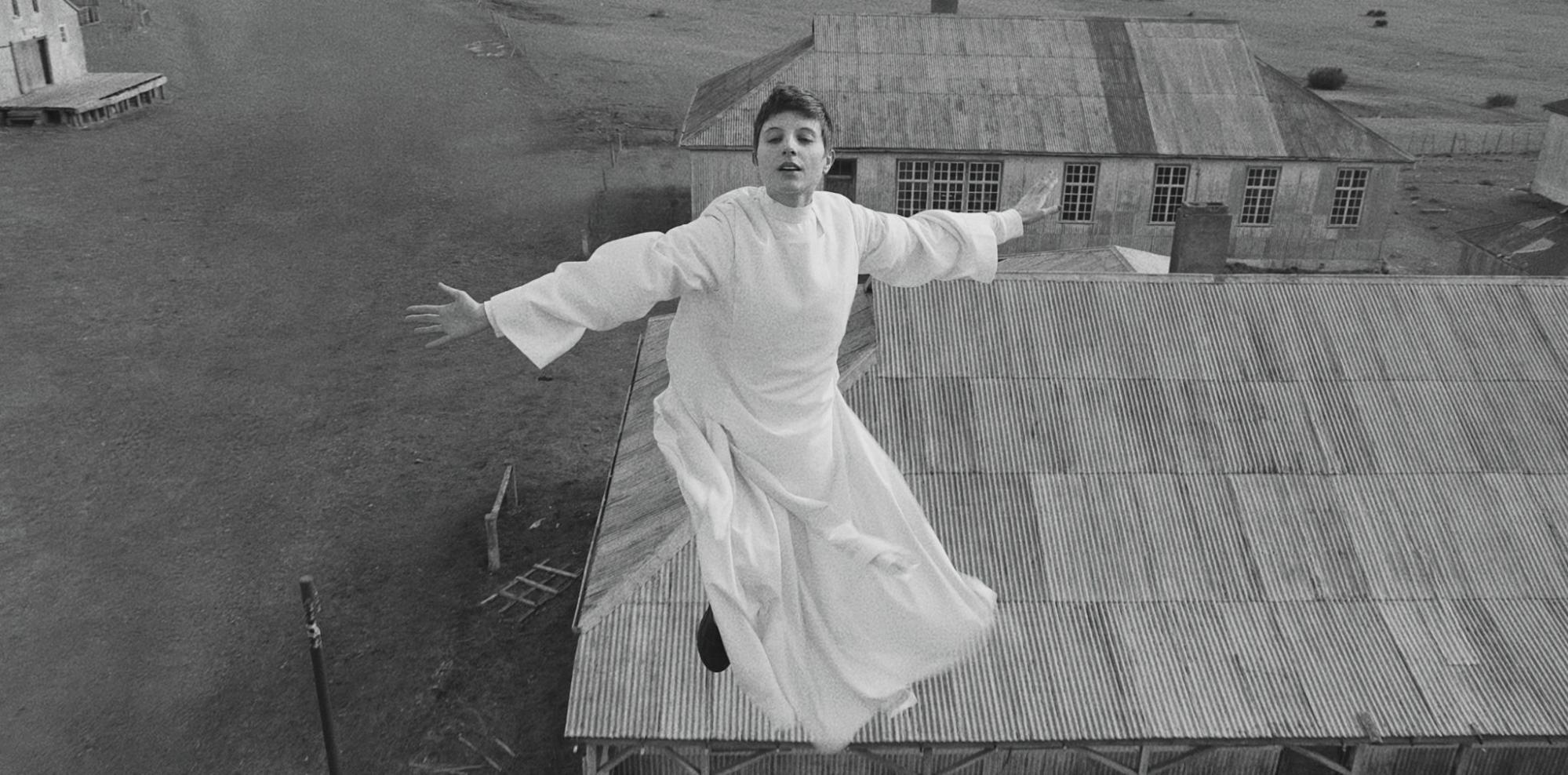 Cena do filme El Conde(2023). A freira Carmen (Paula Luchsinger), está pairando no ar, de braços abertos, ela usa um vestido branco, é uma mulher branca de cabelos curtos, e tem uma expressão de alívio, atrás dela há instalações da fazenda, como um celeiro e galpões