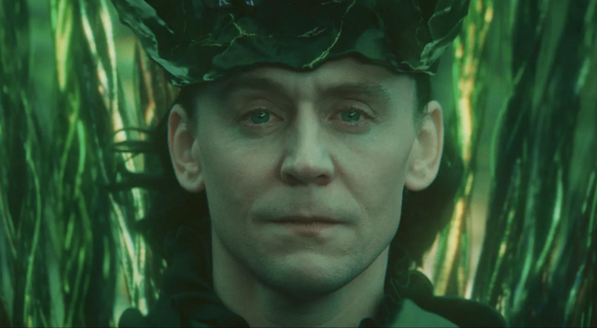 Cena da série Loki. Na imagem, Loki, interpretado por Tom Hiddleston, está olhando para frente, com os olhos marejados e o rosto melancólico. Ele é um homem adulto, de pele branca e olhos claros, cabelos no comprimento dos ombros e de cor escura. Está usando uma coroa na cabeça. A imagem está dando um close-up no rosto de Loki. O fundo é na cor verde escura e toda imagem tem um filtro esverdeado.
