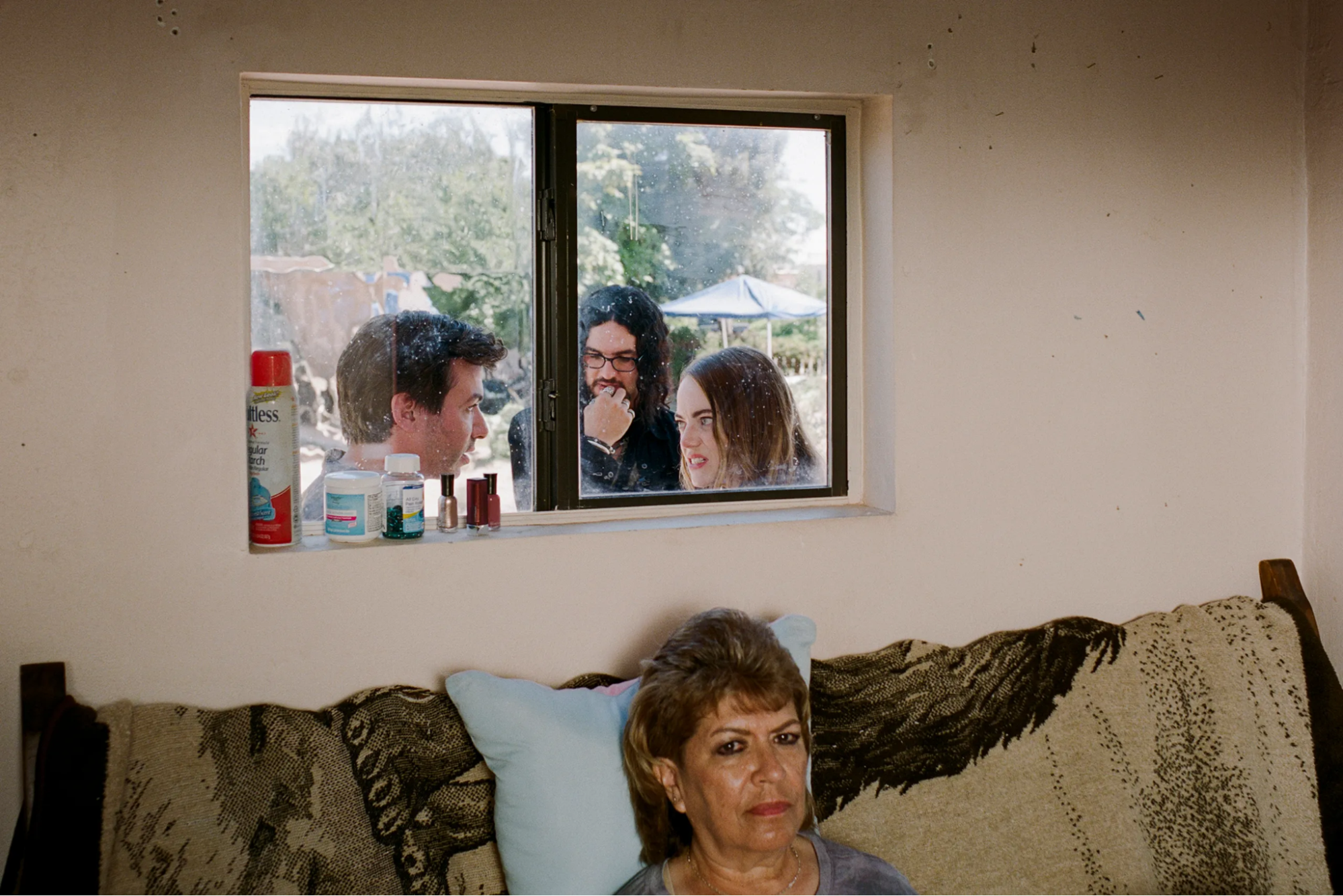 Em primeiro plano, uma senhora, que aparece do pescoço para cima, está sentada em um sofá encostado em uma parede com uma janela logo acima dele. Através da janela, do lado de fora da casa, estão, da esquerda para a direita, os personagens Asher, Dougie e Whitney conversando entre si.