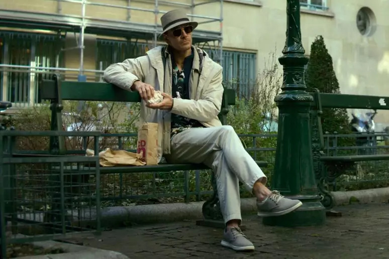 Cena do filme O Assassino. Na imagem, Michael Fassbender está sentado em um banco de praça verde, ao lado de um poste da mesma cor e à frente de um canteiro. Ele é um homem branco, usa calças, casaco e chapéu brancos e óculos escuros. Ele segura um sanduíche.