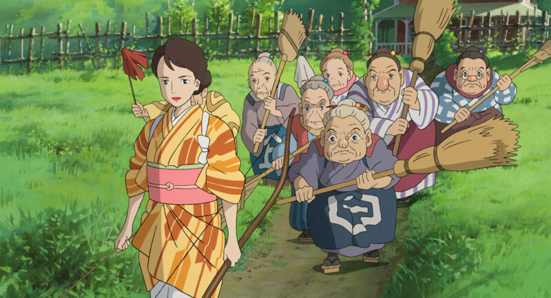 Cena do filme O Menino e a Garça. A cena mostra uma mulher usando um kimono laranja de cabelo marrom e preso. Ela está em primeiro plano e andando. Atrás, seis senhoras estão a seguindo com vassouras na mão e feições irritadas. O ambiente é ao ar livre, um gramado verde, e ao fundo algumas casas atrás da cerca.