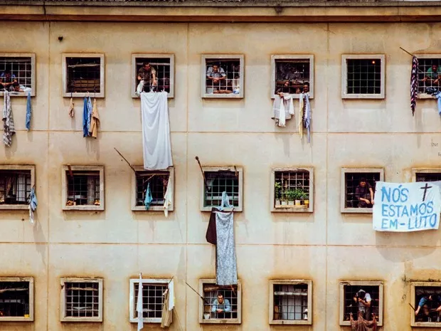 Um prédio amarelado e antigo com muitas janelas e roupas penduradas de forma desorganizada. Presos demonstram luto com cartazes após o Massacre.