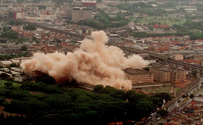 Com vista aérea, é possível ver o Complexo Penitenciário do Carandiru sendo demolido e subindo uma fumaça avermelhada pelo entorno da região.