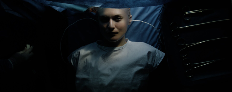 Cena do filme Depois da Morte. Na imagem está uma mulher branca que veste uma camiseta branca lisa. Sua cabeça está raspada e fios colados nela enquanto olha para cima deitada em uma maca de cirurgia com lençóis azuis.