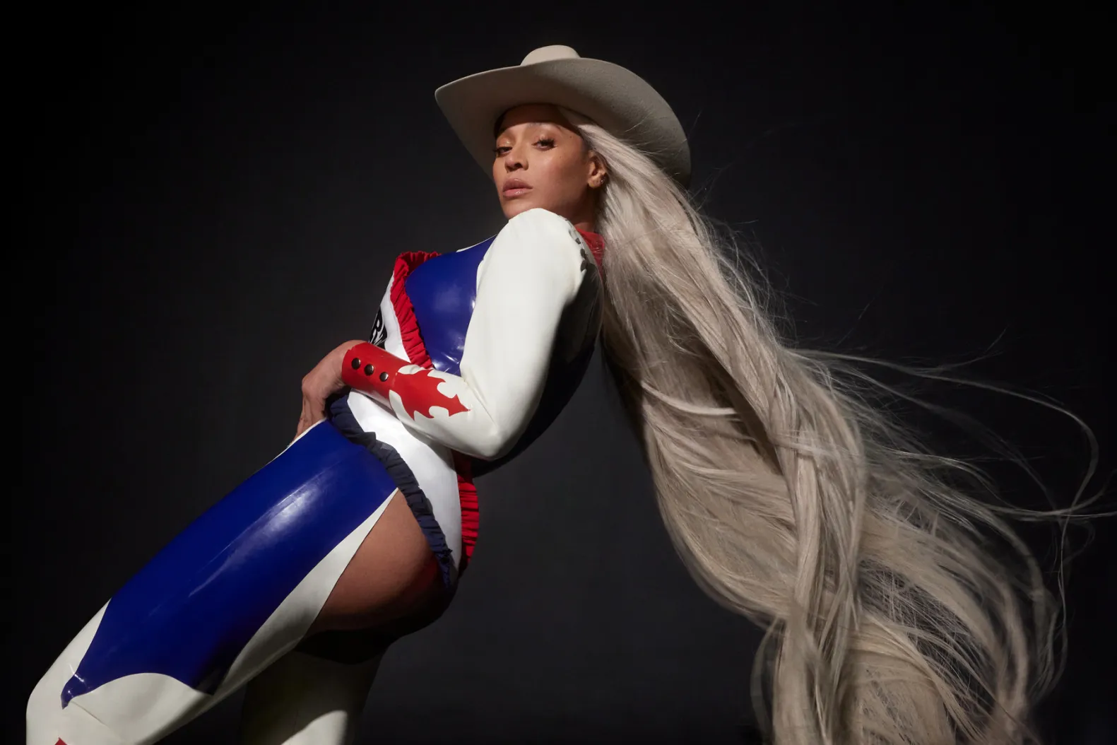 Imagem de Beyoncé para o ensaio fotográfico de COWBOY CARTER. Na fotografia, a artista, uma mulher negra de olhos escuros, aparece com cabelos platinados e longos que vestem um chapéu branco. Ela está de lado para a câmera e veste um chapéu branco com uma vestimenta tradicional de cowboys nas cores branco, azul e vermelho. Ao fundo, o cenário é uma parede cinza que reproduz a sua sombra causada pela iluminação.