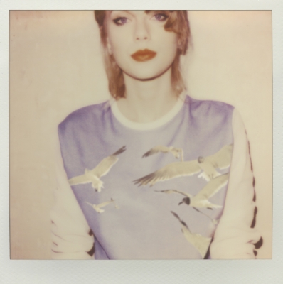 Fotografia quadrada em estética polaroid com uma margem bege. No centro da imagem está Taylor Swift, usando um batom vermelho com um moletom branco e azul com estampas de gaivotas.