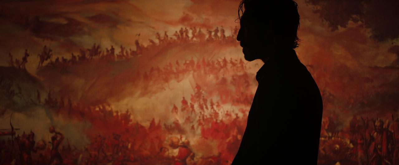 Imagem do filme Fúria Primitiva. Na foto, a sombra do ator Dev Patel está em evidência em frente a uma pintura com as cores marrom e vermelho, que contém diversos guerreiros