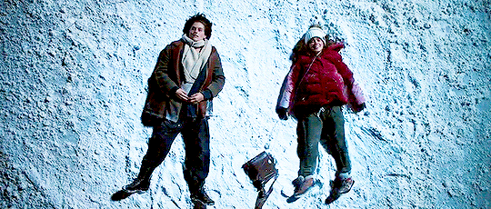 gif de uma cena do filme em que Stella e Will estão deitados na neve. Ambos estão usando roupas de inverno e sondas hospitalares no nariz. Stella abre os braços e pernas deixando marcas na neve. Ambiente escuro, com o foco da iluminação no centro, onde eles estão. Temperatura fria