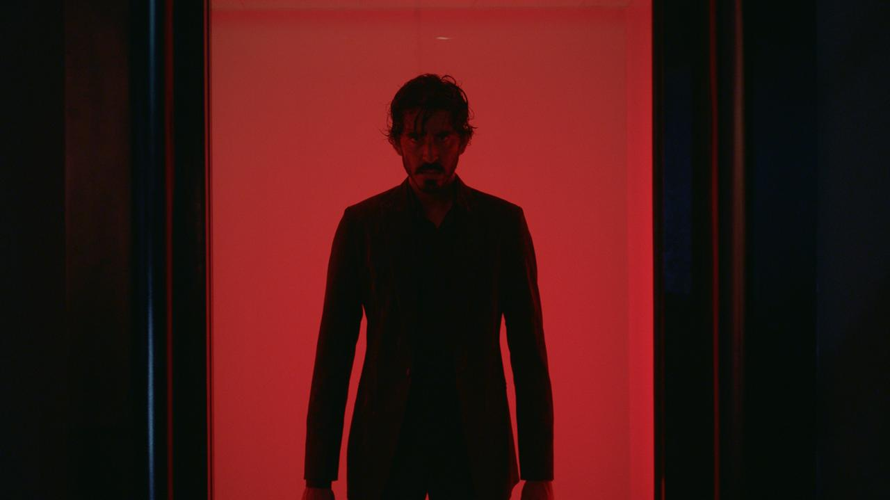 Imagem do filme Fúria Primitiva. Na foto, o ator Dev Patel está dentro de um elevador com um fundo vermelho. Ele está sério e veste uma roupa preta.